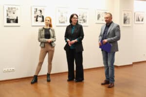 Trzy osoby stoją na tle jasnej ściany, na której wisi rząd czarno-białych fotografii. Od lewej: kobieta o rudych włosach (Dominika Biaduń) w beżowych spodniach i żakiecie, kobieta (Iwona Kędziera) w czarnym garniturze z turkusową bluzką oraz mężczyzna w szarej, wzorzystej marynarce i dżinsach, trzymający niebieską teczkę. Wszyscy patrzą w lewo (poza kadr)