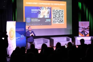 Mężczyzna (Piotr Stec) w granatowej dwurzędowej marynarce i okularach stoi na scenie w świetle reflektorów, gestykuluje w kierunku publiczności. Na dużym ekranie za nim wyświetlony jest slajd podsumowujący z napisem "Dziękujemy i zapraszamy do współpracy", profilem "Społeczeństwo Obywatelskie KPRM" i kodem QR. Na pierwszym planie widać ciemne sylwetki słuchaczy, w tym osobę używające telefonu komórkowego