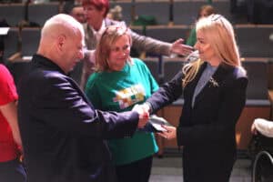 Zbliżenie na moment wręczania dyplomu i gratulacji: kpt. Tomasz Patyra w ciemnym garniturze ściska dłoń Anny Augustyniak w ciemnym kostiumie. Między nimi, w tle, uśmiecha się Sylwia Góźdź w zielonej koszulce, a za nimi widoczna jest widownia z rzędami szarych foteli.