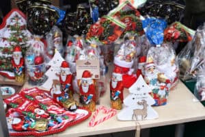 Bliskie ujęcie na asortyment świątecznych słodyczy i drobnych upominków. Na stoisku dominują liczne czekoladowe figurki Świętego Mikołaja w czerwonej, błyszczącej folii, część z nich stoi w świątecznych kubkach. W centrum widoczne są dwa czerwono-białe lizaki w kształcie lasek oraz biała, drewniana ozdoba w kształcie choinki z wyciętym wizerunkiem renifera.