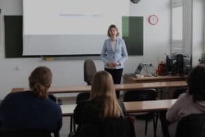 Dorota Krać stoi na środku sali wykładowej przed ekranem rzutnika i patrzy na uczestników widocznych od tyłu. Obok znajduje się biurko z komputerem.