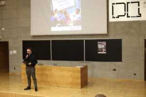 Młody mężczyzna w okularach, ubrany w czarną koszulę i szare spodnie, stoi przed drewnianą katedrą w auli i przemawia do mikrofonu, trzyma w ręku notatnik. W tle znajduje się ściana z szarych płyt betonowych z trzema czarnymi tablicami, na których widnieje plakat wydarzenia. Nad tablicami wisi duży ekran projekcyjny z wyświetloną grafiką „III Edycji Filmoteki empatii”.