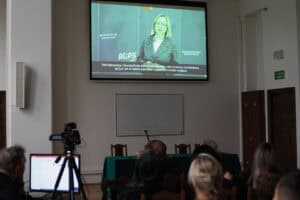 Prezentacja w sali wykładowej, Na dużym ekranie projekcyjnym wyświetlane jest wideo przedstawiające kobietę w szarej marynarce (Małgorzata Romanko), przemawiającą na tle baneru "ROPS Lublin". Na dole ekranu widoczne są napisy (transkrypcja) z treścią: "Taki stereotyp niewątpliwie jest przekazywany, ale możemy powiedzieć, że już nie w takim zakresie, w jakim to miało miejsce.". Na pierwszym planie widać tyły głów publiczności oraz po lewej kamerę wideo na statywie, skierowaną w stronę ekranu