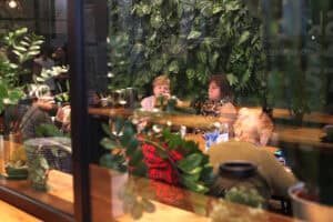 Widok zza przeszklonej ściany na uczestniczki warsztatów siedzące przy drewnianym stole. W centrum kobieta w szarej bluzce i kobieta w beżowym swetrze z panterkowym szalikiem. W tle zielony ogród pionowy, na pierwszym planie liście zamiokulkasa