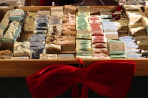 Drewniana skrzynka wypełniona różnokolorowymi, ręcznie robionymi mydłami w kostkach, ułożonymi w rzędach. Kostki mają różne barwy: od błękitu i czerni, przez róż i zieleń, po naturalne beże. Niektóre ozdobione są suszonymi kwiatami lub gwiazdkami anyżu. Z przodu skrzynki zawiązana jest duża, czerwona, aksamitna kokarda.