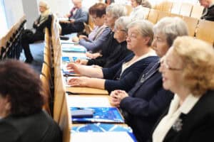 Zbliżenie na uczestników konferencji siedzących w ławkach auli. Widoczny jest profil rzędu starszych osób, głównie kobiet, które z uwagą słuchają wykładu. Na pulpitach przed nimi leżą niebieskie teczki z materiałami konferencyjnymi. Zdjęcie wykonane pod kątem, podkreślające skupienie i zaangażowanie słuchaczy.