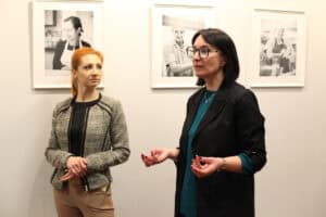 Dwie kobiety stoją na tle wystawy fotograficznej. Kobieta w okularach (Iwona Kędziera) i czarnym garniturze z turkusową bluzką, gestykuluje dłońmi i mówi, patrzy w bok. Obok niej stoi kobieta (Dominika Biaduń) o rudych włosach, w ciemnym żakiecie, która patrzy zasłuchana w bok. W tle widoczne są trzy czarno-białe portrety w białych ramach