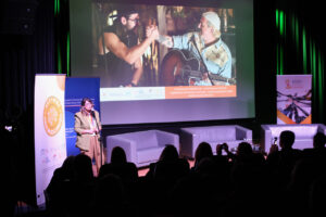 Kobieta (Beata Szewczyk) w beżowym garniturze stoi przy statywie z mikrofonem i przemawia do publiczności. Za nią wyświetlany jest kadr z filmu dokumentalnego, pokazujący dwóch mężczyzn z uściskiem dłoni; jeden z nich trzyma gitarę akustyczną. Pod filmem znajduje się pomarańczowy baner z tekstem opisującym projekt Fundacji EX ORIENTE LUX. Na pierwszym planie widać tyły głów słuchaczy, robiących zdjęcia telefonami.