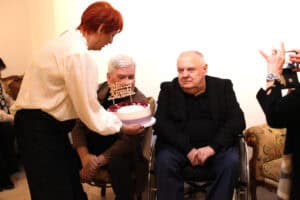 Moment wręczania tortu. Maria Pietrusza-Budzyńska o rudych włosach, ubrana w białą bluzkę, wręcza Januszowi Kirenko siedzącemu na wózku tort z napisem: „Dla Profesora Janusza Kirenki”.