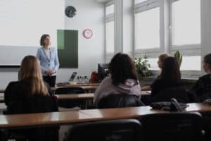 Sala wykładowa, jasno oświetlona światłem wpadającym przez okna z prawej strony. Od lewej Dorota Krać stoi na środku sali. Ma na sobie jasnoniebieski żakiet i czarne spodnie. Na pierwszym planie widoczni są uczestnicy szkolenia, siedzący w rzędach jasnych ławek, widziani od tyłu.