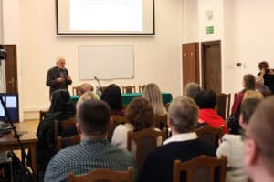 Starszy mężczyzna (prof. Piotr Oleś) z brodą i w okularach, ubrany w ciemną marynarkę, stoi na środku sali wykładowej i przemawia do publiczności, trzyma w ręku mikrofon. Prelegent stoi obok stołu przykrytego zielonym obrusem. Nad nim widoczny jest ekran projekcyjny, a pod ekranem biała tablica. Na pierwszym planie znajdują się tyły głów słuchaczy siedzących na drewnianych krzesłach