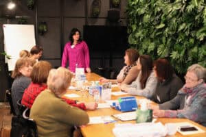 Grupa uczestniczek siedzi przy długim, drewnianym stole warsztatowym. U szczytu stoi prowadząca w koszuli fuksjowej. Na stole leżą materiały szkoleniowe, identyfikatory i tabliczka informacyjna. Prawa ściana pokryta pionowym ogrodem z zielonych roślin.