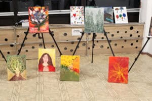 Ekspozycja różnorodnych obrazów namalowanych na płótnie, ustawionych na sztalugach oraz na podłodze w sali. Prace przedstawiają między innymi bukiet kwiatów, portret kobiety z zamkniętymi oczami, las oraz abstrakcyjne słońce.