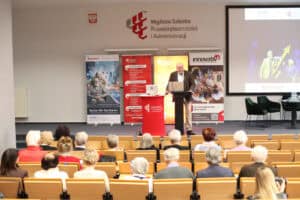 Wystąpienie prelegenta w brązowej marynarce (Wojciech Dec). Mężczyzna stoi przy mównicy w centralnej części. Po lewej stronie sceny stoją cztery pionowe banery, po prawej stronie znajduje się duży ekran multimedialny. Widok z widowni.