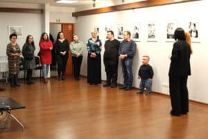 Szeroki kadr przedstawiający grupę około dziesięciu osób, w tym jednego niskorosłego mężczyznę w dżinsach, stojących w rzędzie na drewnianej podłodze w sali wystawowej. Zwróceni są w stronę kobiety w czarnym garniturze, która stoi tyłem do widza. W tle wisi rząd czarno-białych fotografii w białych ramach