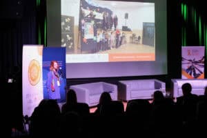 Kobieta z rudymi włosami (Magdalena Kara-Sajek), ubrana w niebieską tunikę i kolorową apaszkę, stoi przy statywie z mikrofonem i wygłasza prelekcję. Na ekranie w tle wyświetlany jest slajd z wizerunkiem dzieci stojących w rzędzie podczas wydarzenia artystycznego. Pod nim widnieje napis "Stowarzyszenie WSPAK - Sztuka Daje Siłę". Na pierwszym planie widoczne są tyły głów słuchaczy siedzących w zaciemnionej sali