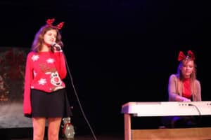 Na ciemnej scenie, oświetlonej lekko niebieskim światłem, stoi młoda dziewczyna z długimi, brązowymi włosami, ubrana w czerwony sweter ze wzorem renifera i czarną spódniczkę. Ma na głowie opaskę z czerwonymi porożami renifera i śpiewa do mikrofonu. Po jej prawej stronie, przy białym keyboardzie, siedzi kobieta w szarym kardiganie, czerwonym topie i dużej, czerwonej kokardzie na opasce.