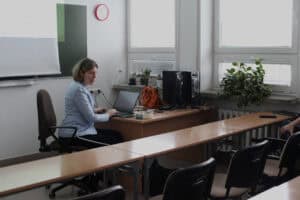 Dorota Krać siedzi przy biurku ustawionym pod oknem w sali wykładowej i pracuje na laptopie. Na biurku stoi monitor i komputer stacjonarny, obok leży torebka