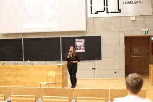 dr inż. Agnieszka Gandzel w okularach i ciemnym stroju stoi w auli wykładowej przed drewnianą katedrą i przemawia do uczestników, używa mikrofonu. W tle widoczna jest ściana z szarych płyt betonowych, na której zamontowano czarne tablice. Na jednej z tablic przyczepiony jest plakat wydarzenia „III Edycja Filmoteki empatii”. Nad tablicami znajduje się fragment dużego ekranu projekcyjnego.