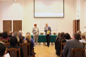 Dwoje prelegentów stoi w sali wykładowej: kobieta o rudych włosach (Maria Pietrusza-Budzyńska) w jasnym, kraciastym garniturze i uśmiechnięty mężczyzna (prof. Wojciech Otrębski) w szarym garniturze, trzymający mikrofon. Za nimi widoczny jest ekran projekcyjny z tytułem "KONFERENCJA NAUKOWO-SZKOLENIOWA" i datą "20. 11. 2025". Uczestnicy, siedzą na drewnianych krzesłach w rzędach, obserwują prelegentów. W tle widoczne są również ciemne, drewniane drzwi i szafy