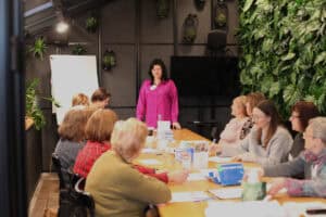 Sala warsztatowa z kilkunastoma uczestniczkami siedzącymi przy długim stole. Prowadząca w koszuli fuksjowej stoi za stołem, uśmiecha się. Na stole rozłożone materiały, długopisy, identyfikatory i tabliczka projektu. Tło: czarna metalowa ściana z wiszącymi roślinami oraz ogród pionowy po prawej.