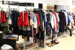 Dwa długie wieszaki (stojaki) wypełnione kolorowymi ubraniami, przygotowane w ramach akcji wymiany odzieży. Na wieszakach widoczne są m.in. czerwone spódnice, ciemne płaszcze, kolorowe sukienki i swetry. Pod wieszakami na podłodze ustawionych jest kilka par butów.