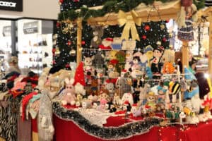 Bogato wyposażone stoisko z rękodziełem w centrum handlowym. Drewniana konstrukcja stoiska ozdobiona jest zieloną girlandą, złotą gwiazdą i lampkami. Na czerwonym obrusie i półkach wyeksponowano dziesiątki kolorowych, dzierganych zabawek, maskotek oraz zimowych ubrań. W tle widać dużą choinkę i witryny sklepowe.