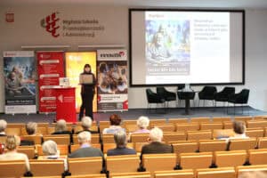 Na scenie, przy mównicy, stoi kobieta (Katarzyna Barcikowska) w ciemnym stroju. Na dużym ekranie obok niej wyświetlany jest slajd z napisem: „Nasza Misja: Budujemy Lublin bez barier, w którym głos seniorów jest słyszany i ważny”. Slajd zawiera również zdjęcie uśmiechniętej pary seniorów oraz logo projektu „Senior 60+ bez barier”. Widok z widowni.