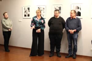 Cztery osoby stoją na drewnianej podłodze w galerii. Kobieta (Agnieszka Różyło) w kwiecistej bluzce i czarnych szerokich spodniach, stoi przodem i gestykuluje dłońmi, mówi do kogoś poza kadrem. Obok niej (po lewej i prawej) stoją: kobieta w szarej kurtce oraz dwóch mężczyzn, z których jeden jest w czarnym t-shircie, a drugi w granatowym swetrze. W tle wiszą cztery czarno-białe portrety