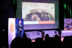 Od lewej mężczyzna (Mateusz Popławski) w ciemnym garniturze i krawacie stoi na scenie i przemawia do publiczności. Na dużym ekranie za nim wyświetlone jest zdjęcie z perspektywy tunelu, w którym tłoczą się ludzie, oraz podpis "Łukowskie Forum Rozwoju - Łukowski Spacerownik 2025". Na pierwszym planie widać tyły głów publiczności siedzącej w zaciemnionej sali