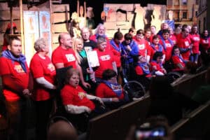 Grupowe zdjęcie uczestników wydarzenia na scenie. Większość osób ma czerwone koszulki z logo i niebieskie chusty na szyi; część siedzi na wózkach inwalidzkich na pierwszym planie. W centrum stoją mężczyzna w czarnej koszulce oraz Anna Augustyniak trzymająca dokument. W tle znajduje się bogata drewniana scenografia z domkami, rzeźbami w ludowych strojach i malowanymi postaciami świętych. Grupa jest liczna i uśmiechnięta.