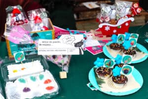 Fragment stoiska Wydziału Historii i Archeologii. W przezroczystym opakowaniu na pierwszym planie znajdują się kolorowe pianki galaretkowe nabite na wykałaczki. Po prawej stronie, na dwóch turkusowych, plastikowych talerzykach, leżą ciemne babeczki z polewą czekoladową, ozdobione niebieskimi gwiazdkami i papierowymi pikami z logo UMCS.