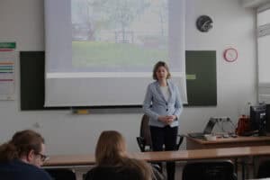 Dorota Krać w jasnoniebieskim żakiecie stoi przed grupą i prowadzi wykład. W tle, na dużym ekranie rzutnika, wyświetlany jest kadr z obrazem krajobrazu: trawiasty teren, drzewa i drewniany sprzęt rekreacyjny przy wodzie (huśtawka). Po bokach ekranu wiszą ciemnozielone tablice.