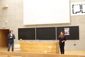 Aula wykładowa Politechniki Lubelskiej. Po prawej stronie, przed drewnianą katedrą, stoi dr inż. Agnieszka Gandzel w ciemnym stroju, przemawia do mikrofonu. Po lewej stronie kadru stoi dr hab. Andrzej Kapusta w okularach i ciemnej marynarce, trzyma w dłoniach mikrofon i telefon. W tle widoczna jest ściana z szarych płyt betonowych z trzema czarnymi tablicami, na których zawieszono plakat „Filmoteki empatii”. Nad tablicami znajduje się duży, biały ekran projekcyjny, a po prawej stronie, u góry, czarny logotyp uczelni.