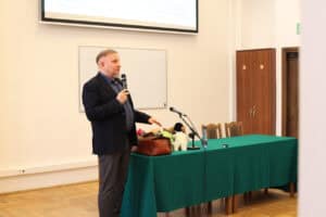 Mężczyzna (dr Krzysztof Ciepliński), w ciemnej marynarce i szarych spodniach stoi z boku stołu przykrytego długim, zielonym obrusem, trzyma w ręku mikrofon i gestykuluje w trakcie przemówienia. Na stole widoczne są brązowa skórzana torba, maskotka przypominająca pingwina oraz statyw mikrofonowy. Za prelegentem widać białą tablicę i ekran projekcyjny z niewyraźnym tekstem, a w tle szafę i drzwi