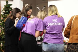 Grupa kobiet pochylona nad stołem podczas kiermaszu. Dwie kobiety na pierwszym planie mają na sobie fioletowe koszulki z białym napisem „Moc Dobrostanu”. W tle po lewej stronie widać udekorowaną choinkę.