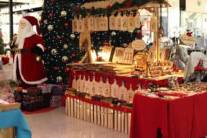 Stoisko z wyrobami z drewna pod drewnianym dachem ozdobionym igliwiem. Na stoisku i pod nim zawieszone są liczne deski do krojenia z wypalanymi grafikami i napisami. Obok stoiska stoi duża figura Świętego Mikołaja z latarnią, otoczona prezentami pod wielką choinką.