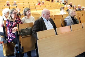 Uczestnicy konferencji siedzą w rzędach auli, patrzą w stronę sceny. W centrum kadru siedzi mężczyzna w brązowej marynarce (Wojciech Dec). Obok niego siedzą dwie kobiety – jedna w kolorowej bluzce i okularach (Justyna Spryszak) i druga w jasnej dzianinowej bluzce (Dorota Krać). W tle widać kolejne rzędy zajęte przez uczestników.