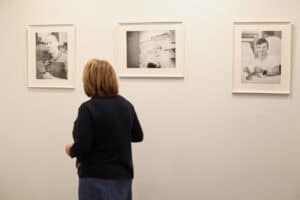 Widok na trzy czarno-białe fotografie w białych ramach, wiszące na jasnej ścianie. Kobieta o blond włosach, ubrana w ciemną bluzkę i niebieską spódnicę, stoi tyłem do widza i ogląda środkowe zdjęcie. Na fotografiach widoczne są portrety pracujących osób