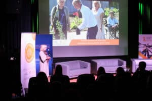 Od lewej kobieta (Beata Zawadzka) o jasnych włosach, ubrana w jasną sukienkę i szal, stoi przy mikrofonie i przemawia do publiczności. Na ekranie w tle wyświetlony jest kadr ze slajdu lub filmu, przedstawiający cztery osoby, w tym mężczyznę sadzącego roślinę i kobietę na wózku inwalidzkim. Pod obrazem widnieje napis "Fundacja Gdzieś na Roztoczu – Integracja mieszkańców Gminy Goraj". Na pierwszym planie widać ciemne sylwetki publiczności.