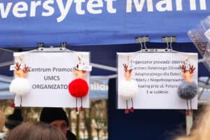 Zadaszenie niebieskiego namiotu z widocznym białym napisem. Na dwóch wiszących arkuszach papieru, przyczepionych klipsami biurowymi, widnieje informacja: „Centrum Promocji UMCS ORGANIZATOR" oraz: „Organizator prowadzi zbiórkę na rzecz Dziennego Ośrodka Adaptacyjnego dla Dzieci i Młodzieży z Niepełnosprawnością Intelektualną przy ulicy Poturzyńskiej 1 w Lublinie”. Do arkuszy doczepione są breloki przedstawiające puszyste renifery w kolorach białym, czerwonym i szarym z czerwonymi butami.
