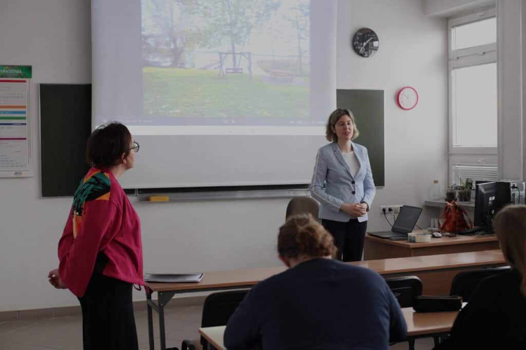 Dwie wykładowczynie - Anna Bieganowska-Skóra i Dorota Krać stoją przed uczestnikami wykładu. W tle na rzutniku wyświetlany jest kadr krajobrazowy (trawa, drzewa, drewniane sprzęty nad wodą).