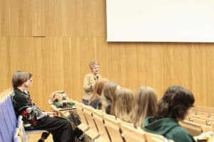 prof. Halina Rarot, w jasnobrązowej marynarce, stoi w przejściu auli wykładowej, przemawia do mikrofonu. Jest zwrócona w stronę słuchaczy, którzy siedzą w ławach z jasnego drewna oraz na niebieskich krzesłach. Tło stanowi wysoka ściana wyłożona pionowymi panelami drewnianymi, nad którą widoczny jest fragment dużego, białego ekranu projekcyjnego.