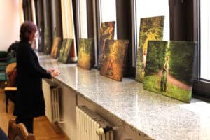 Kobieta w ciemnym stroju z krótkimi, brązowymi włosami stoi przy długim, szerokim parapecie, na którym ustawiono rząd pionowych i poziomych wydruków na płótnie. Wszystkie przedstawiają motywy leśne, a dwa najbliższe ukazują postać kobiety w stroju stylizowanym na naturę. Parapet ma szaro-białą, nakrapianą teksturę, pod nim widoczny jest grzejnik, a w tle duże okna