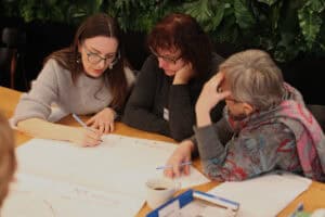 Trzy kobiety pochylone nad arkuszem papieru. Po lewej w szarym swetrze pisze długopisem, w środku kobieta w ciemnym swetrze opiera głowę na dłoni, po prawej kobieta w kolorowej chuście trzyma rękę przy czole. W tle fragment ściany z zielonych roślin.