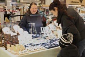 Uśmiechnięta wystawczyni za stoiskiem z rękodzielniczą biżuterią. Na jasnym blacie, doświetlonym lampami, wyeksponowano liczne naszyjniki na białych popiersiach oraz kolczyki na drewnianych ekspozytorach. Biżuterii przygląda się klientka z dzieckiem w zimowej czapce.