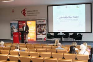 Prelegent w brązowej marynarce (Wojciech Dec) stoi przy czarnej mównicy, patrząc w ekran laptopa. Na monitorze za jego plecami widnieje slajd z tytułem: „Lubelskie Bez Barier. Strategia Rozwoju Lubelskiego Partnerstwa na Rzecz Dostępności na lata 2025-2028”. Tło slajdu jest jasne z delikatnym konturem architektury Lublina. Widok z widowni