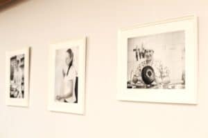 Na jasnej ścianie wiszą trzy czarno-białe fotografie w białych ramach, ustawione są perspektywicznie, od małej do dużej. Największe, znajdujące się na pierwszym planie, przedstawia uśmiechniętą kobietę w czepku, trzymającą garnek i pracującą w kuchni, na tle wiszących noży. Dwa pozostałe zdjęcia w tle są nieostre