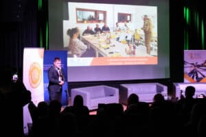 Mężczyzna (Grzegorz Chmielewski) w ciemnym garniturze, stoi na scenie przy mikrofonie, z dłońmi splecionymi na brzuchu, przemawia. Na ekranie w tle wyświetlony jest slajd przedstawiający spotkanie przy długim stole w pomieszczeniu, przy którym siedzi grupa osób, a przed nimi stoi wojskowy. Pod slajdem widnieje napis "Lubelskie Stowarzyszenie Ekonomii Społecznej - Gotowi na Kryzys". Na pierwszym planie widać publiczność, w tym osobę klaszczącą, w dalszej części kadru trzy szare sofy