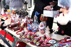 Stół na kiermaszu, pełen słodyczy i dekoracji. W centrum widoczne są ręce: jedna trzyma szare prostokątne pudełko, a druga wrzuca w nie banknot 10 zł. Na stole leżą czekoladowe figurki Mikołajów owinięte w czerwoną i złotą folię, lizaki laski oraz ozdoby.